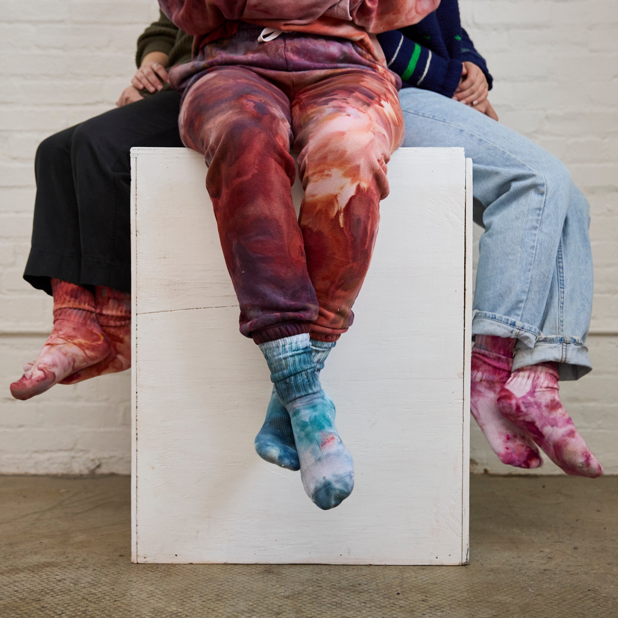 Three pairs of Goldy Way's hand-dyed crew socks, each pair on the feet of a model. From left to right, colors shown are Solstice (orange with reds and purple), Lake Effect (blue with darker blue and pink), and Winterberry (dark fuchsia with blue and orange).