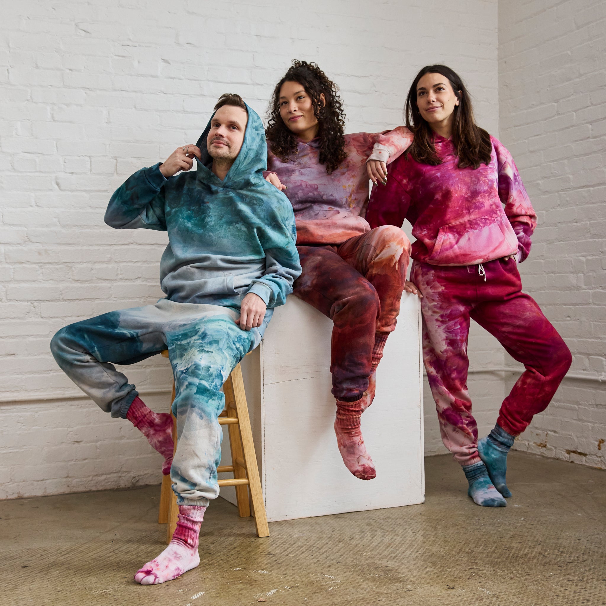 Goldy Way's hand-dyed crew socks and sweatsuits, worn by three models, each wearing a different color. From left to right, models are wearing the Lake Effect sweatsuit with Winterberry socks, the Solstice sweatsuit with Solstice socks, and the Winterberry sweatsuit with Lake Effect socks. 