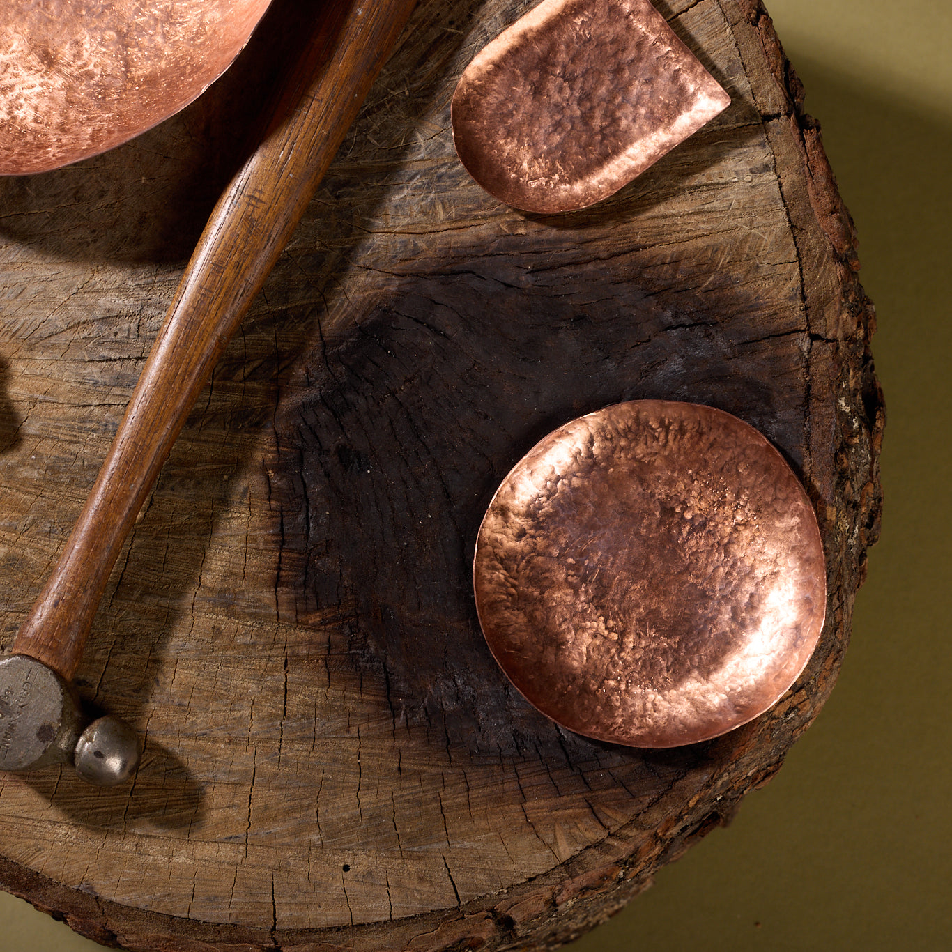 Hammered brass trinket trays sit on a stump. There is a hammer with a wooden handle for working brass next to the trays.