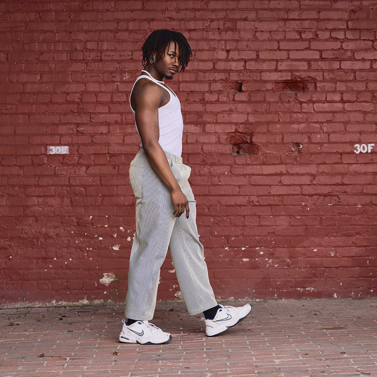 Person wearing a white tank top and gray pants standing against a red brick wall.