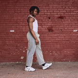 Person wearing a white tank top and gray pants standing against a red brick wall.