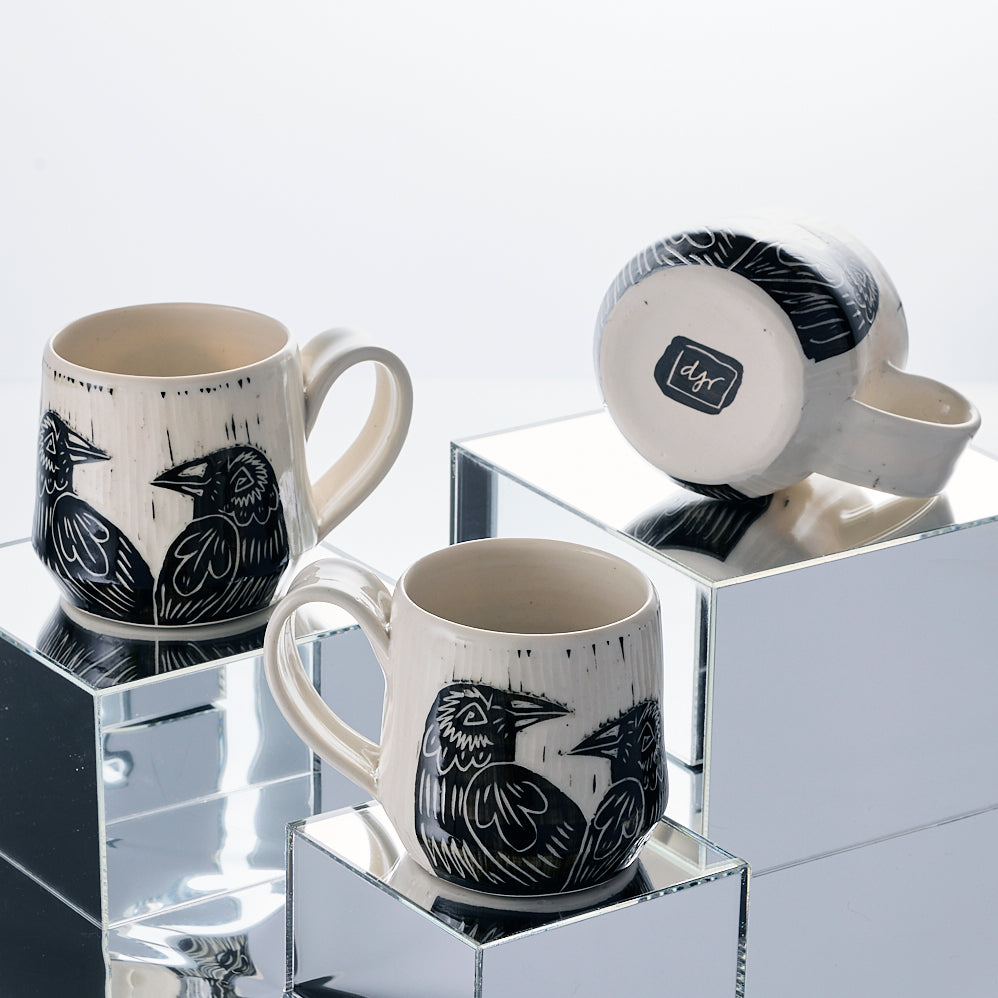 Three ceramic mugs with raven  illustrations on a reflective surface PostbyDJR