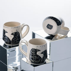 Three ceramic mugs with raven  illustrations on a reflective surface PostbyDJR