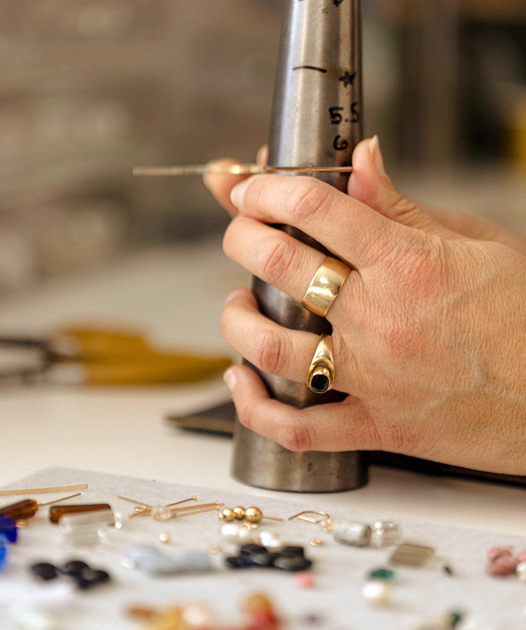 Hand holding a metal tool over a jewelry-making setup with beads and tools on a table.
