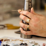 Hand holding a metal tool over a jewelry-making setup with beads and tools on a table.